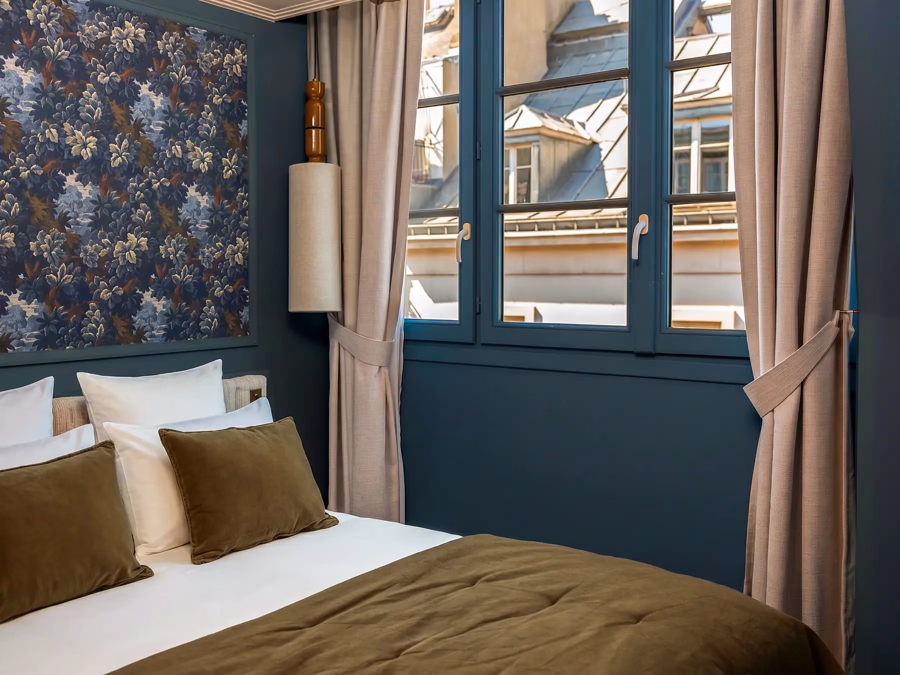 Chambre bleu marine avec tapisserie fleurie, lit couvert d'un jeté kaki, fenêtre sur façade parisienne.