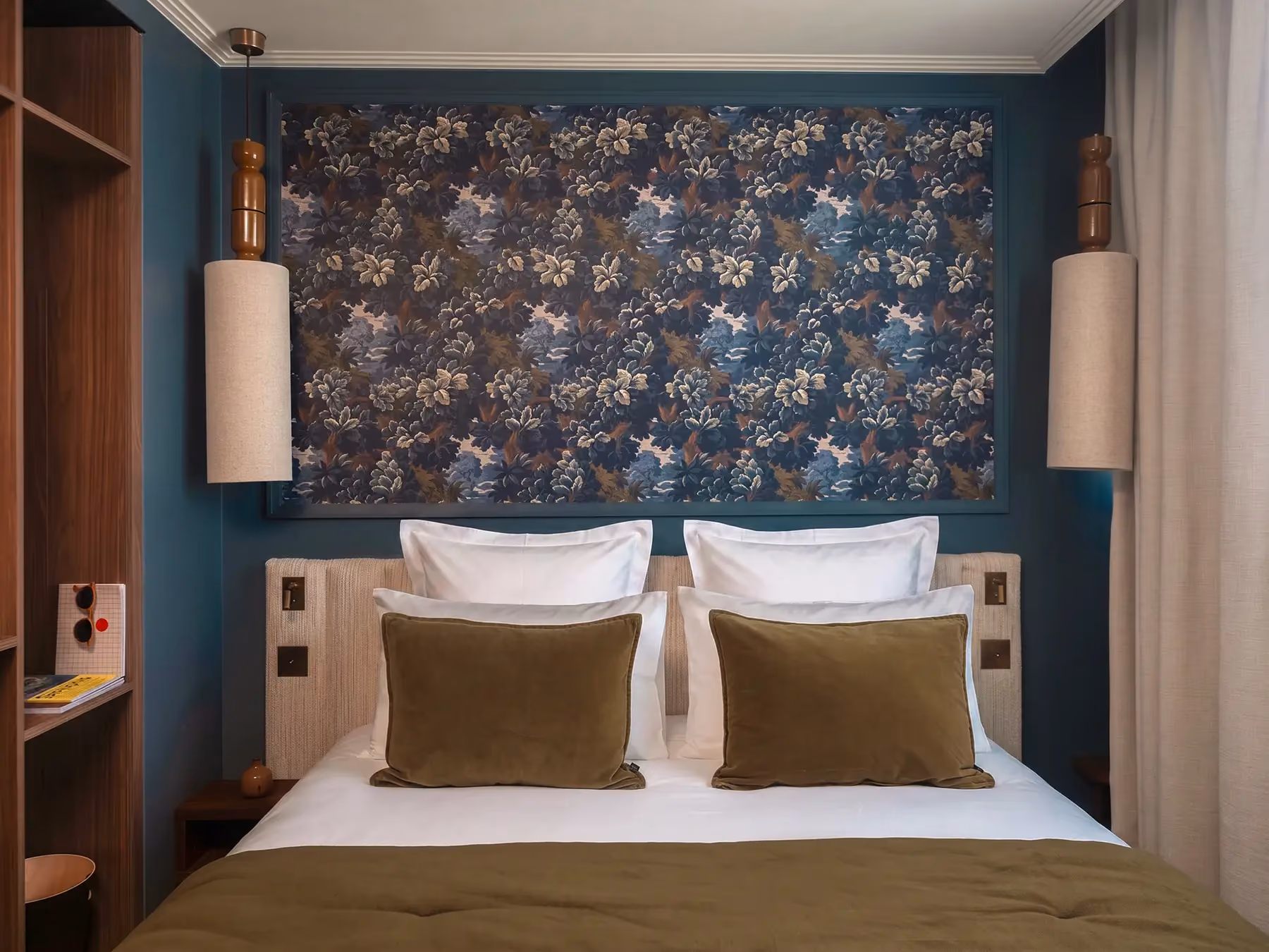 Tête de lit avec papier peint bleu fleuri et coussins kaki, deux suspensions cylindriques.