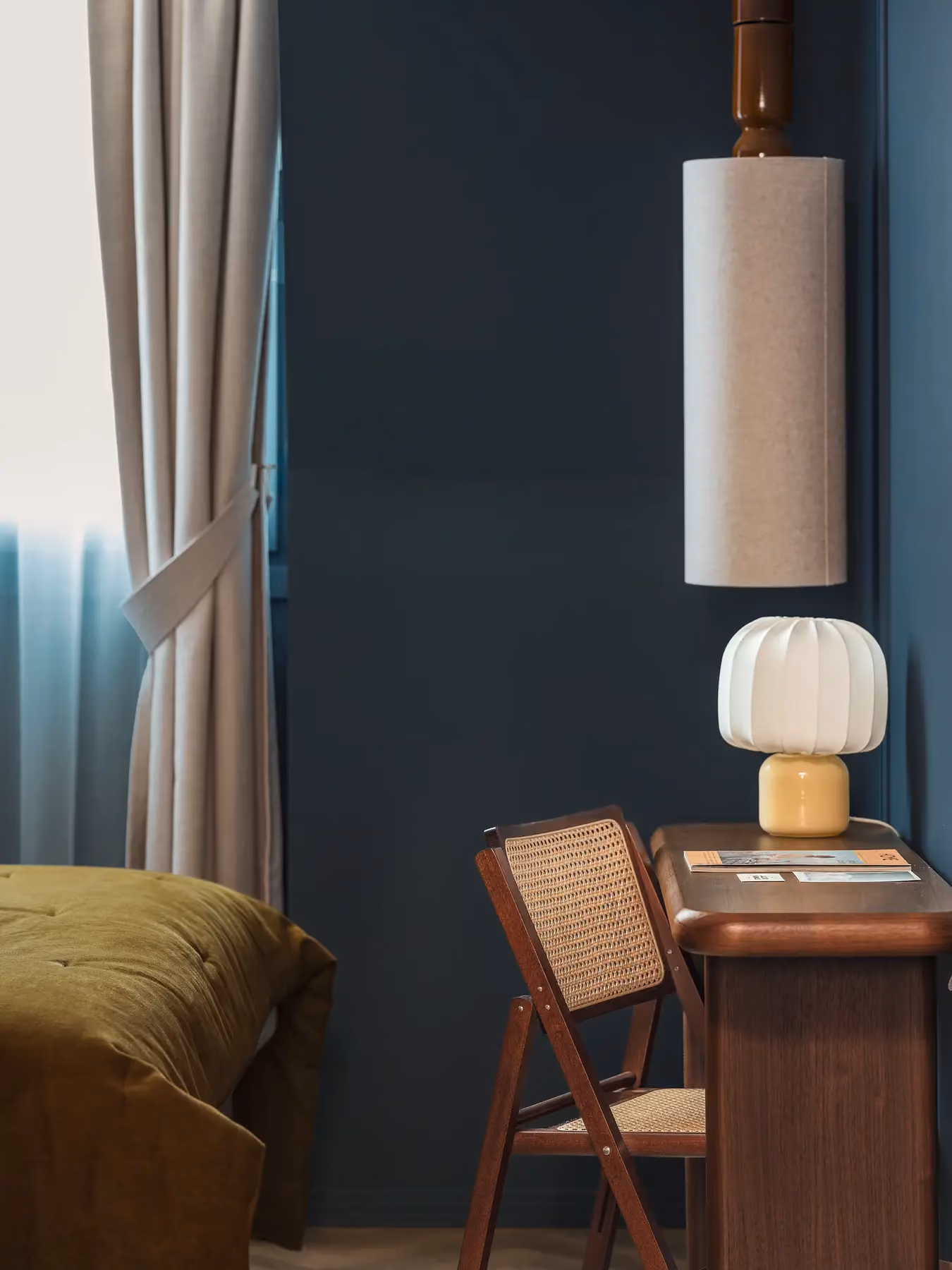 Bureau en bois avec lampe champignon et chaise cannée près d'une fenêtre voilée, mur bleu profond.
