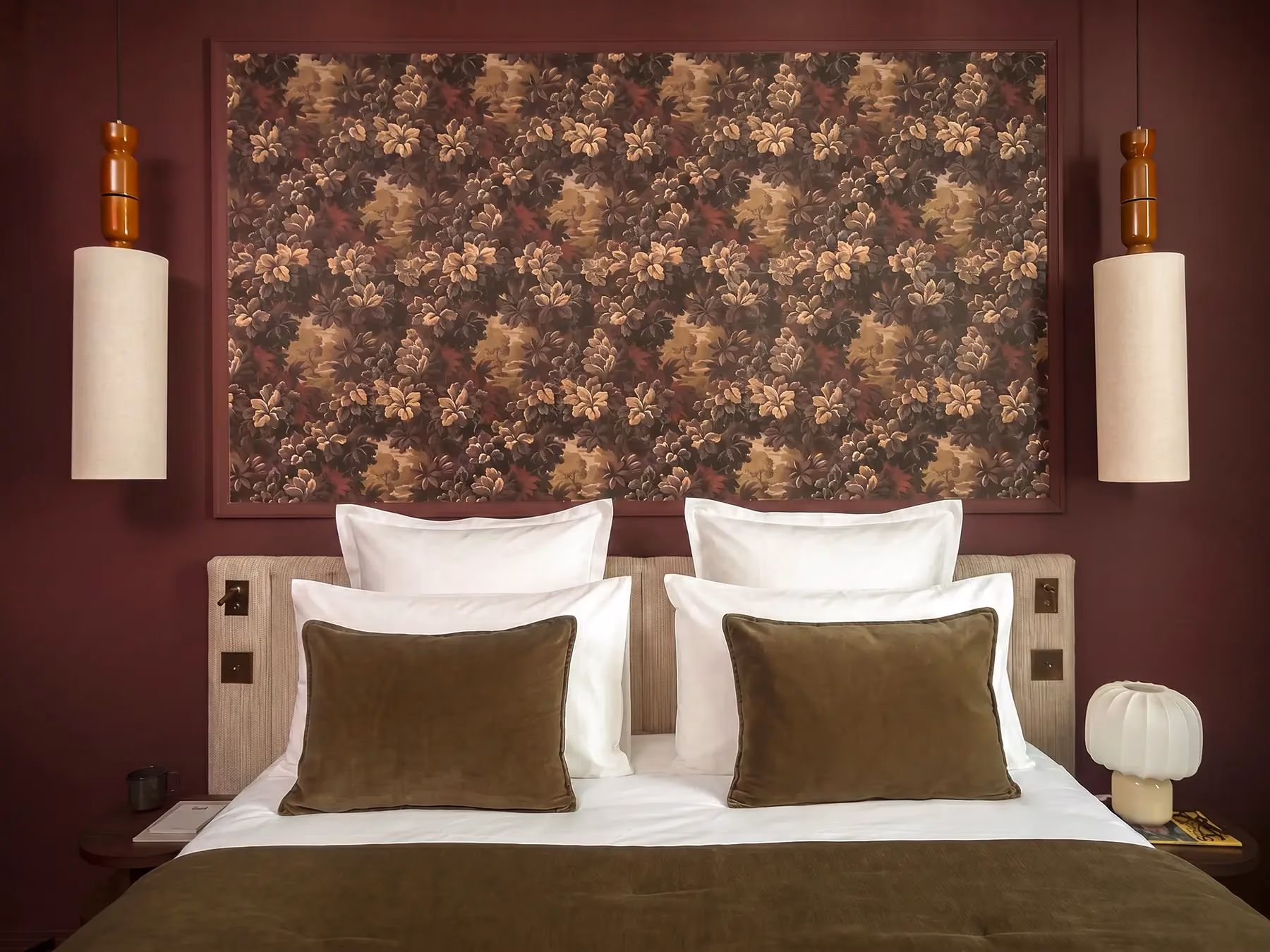 Tête de lit avec tapisserie florale, coussins vert olive et appliques cylindriques.