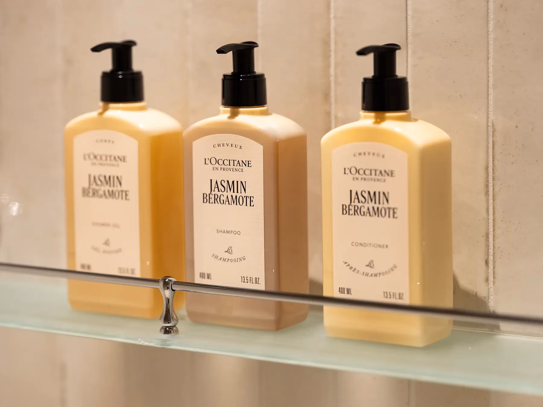 Trio de flacons L'Occitane "Jasmin Bergamote" (douche, shampooing, après-shampooing) sur étagère en verre.