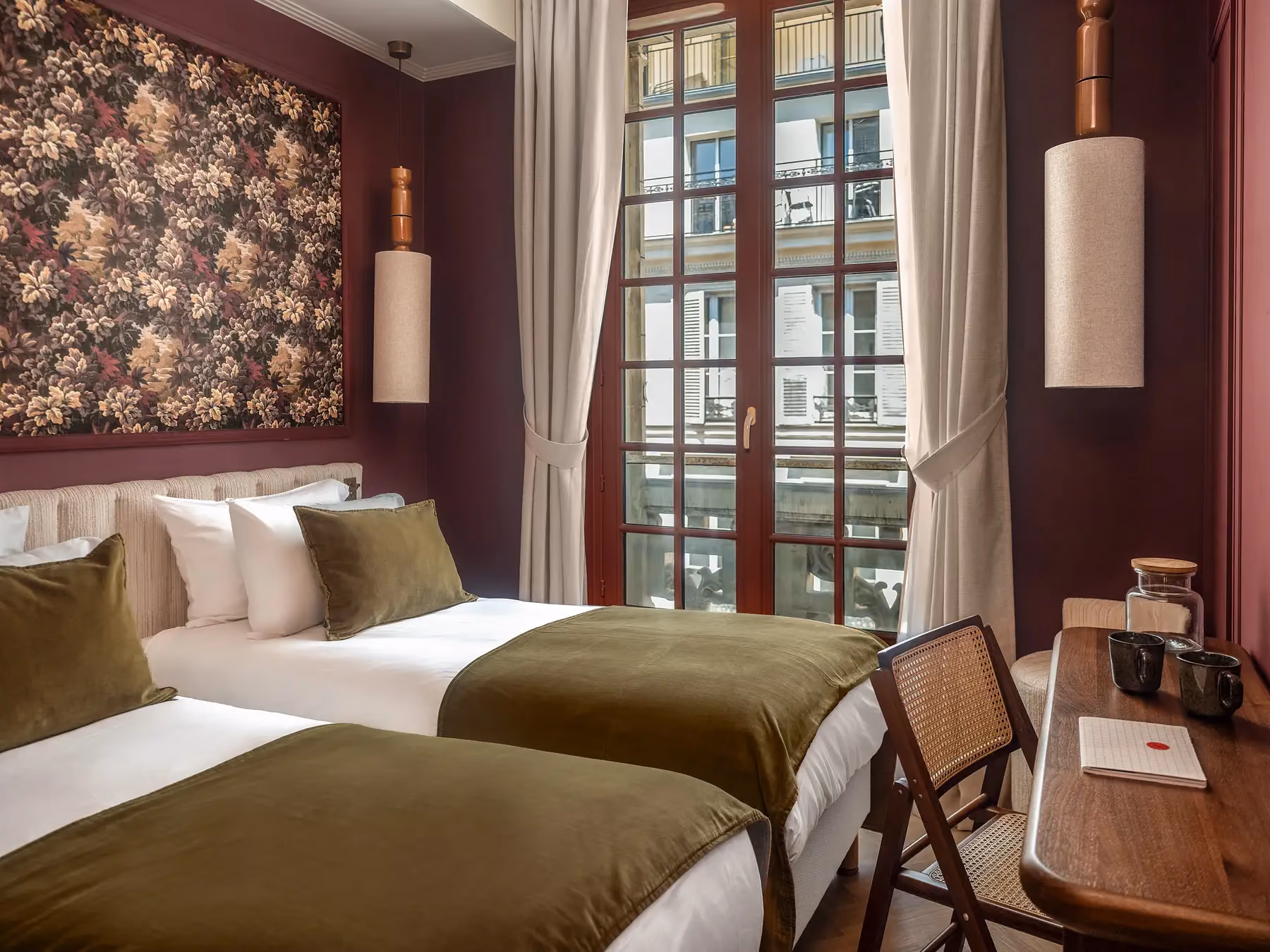 Chambre d'hôtel bordeaux avec lits jumeaux, jetés vert olive et fenêtre à balcon parisien.
