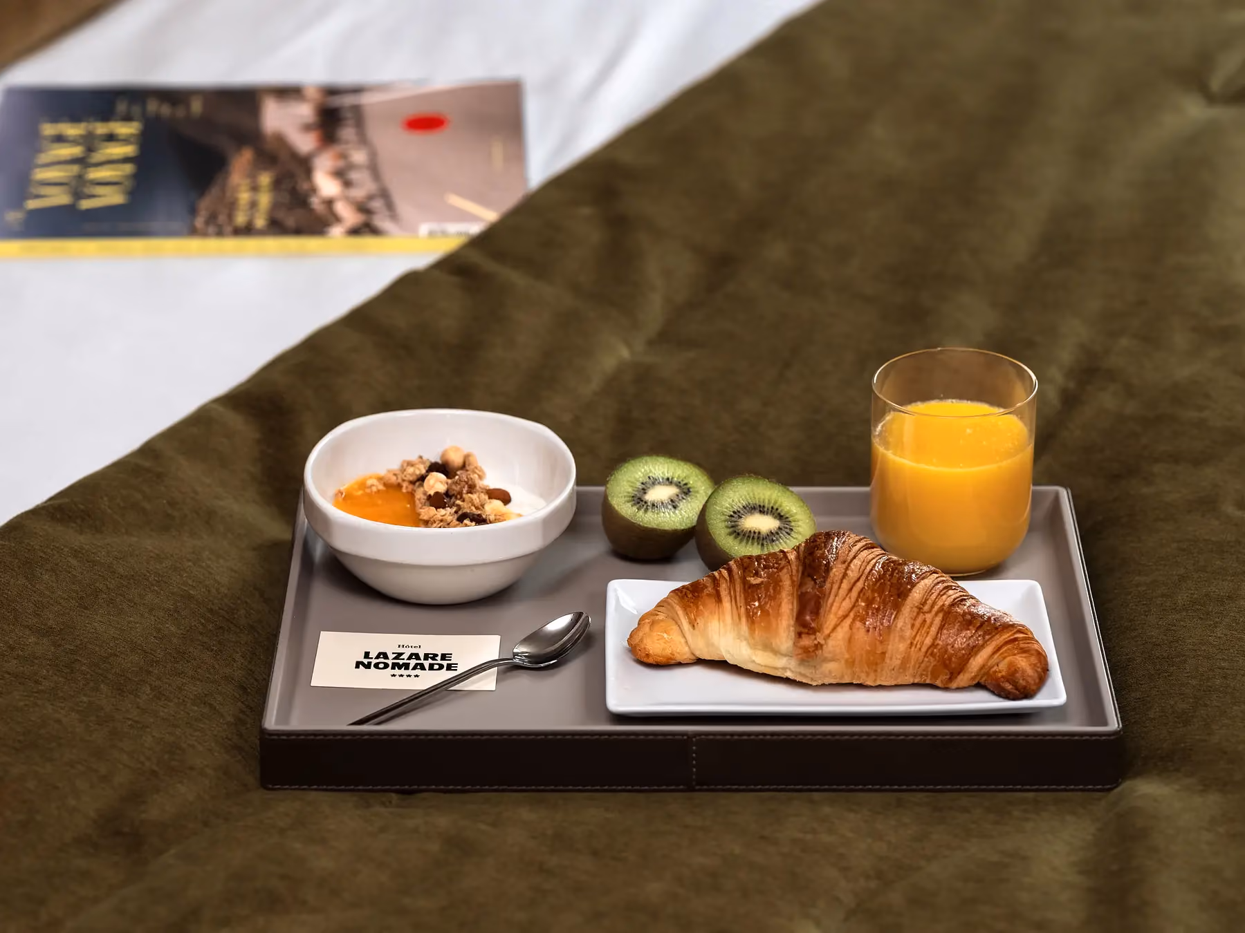 Plateau de petit-déjeuner "Hôtel Lazare Nomade" avec croissant, kiwi, granola et jus d'orange sur un lit.Plateau petit-déjeuner disposé sur un lit, composé d'un croissant, d'un bol de muesli, d'un kiwi, d'un verre de jus d'orange et d'une carte au nom de l