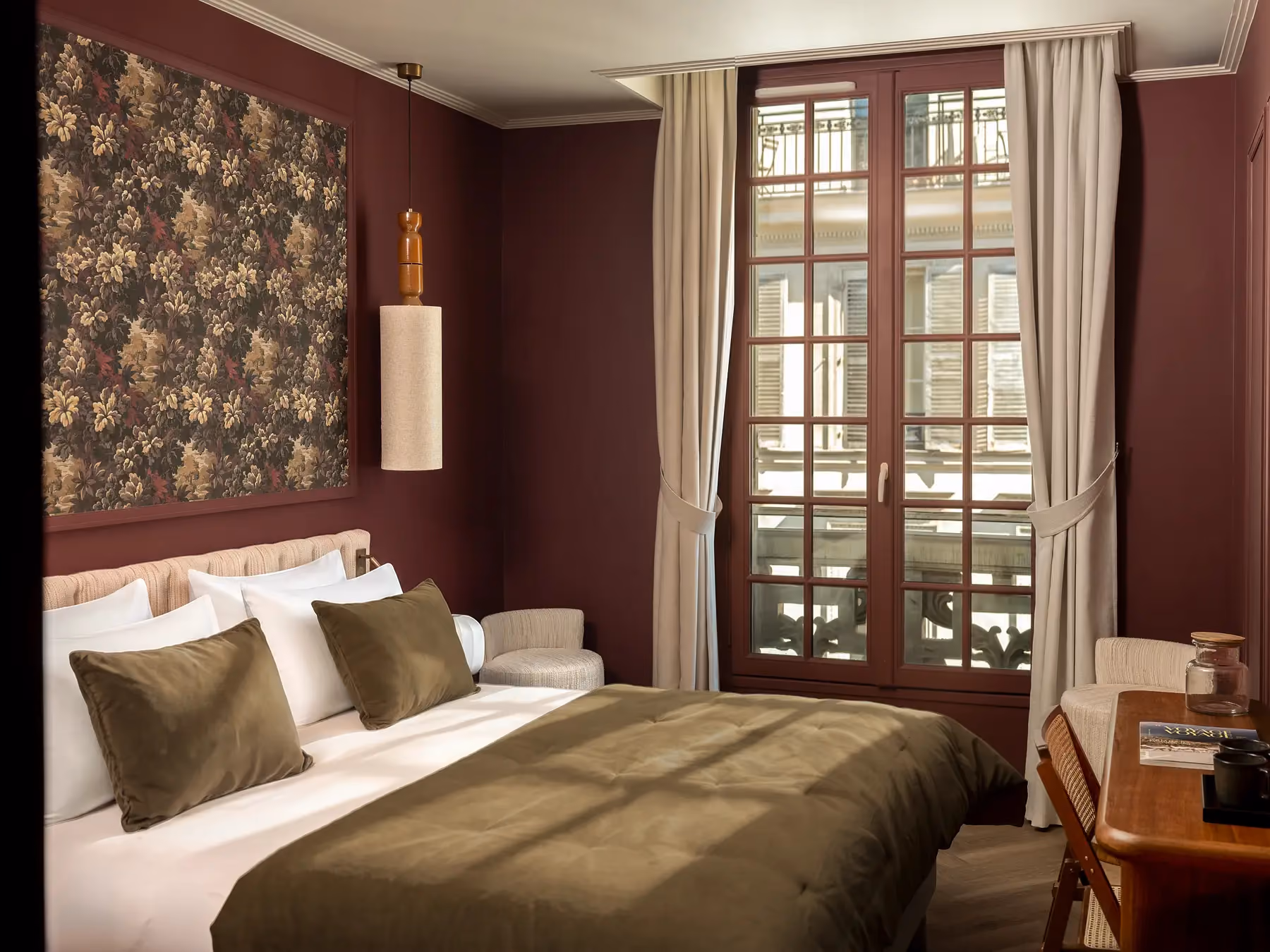 Chambre bordeaux avec lit à jeté olive, grande fenêtre ouvragée et bureau en bois.