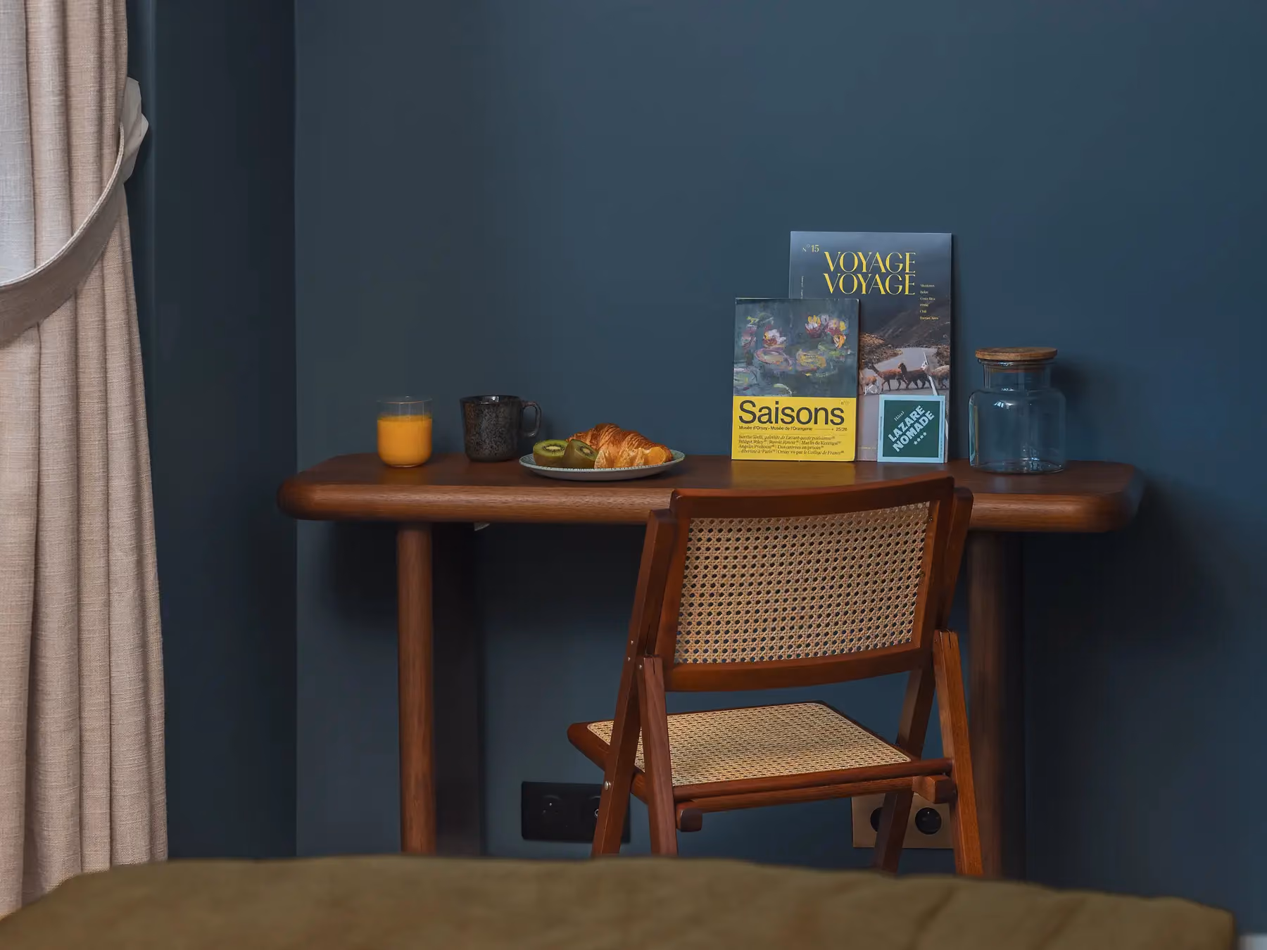Bureau en bois avec chaise cannée près d'une fenêtre voilée, mur bleu foncé.