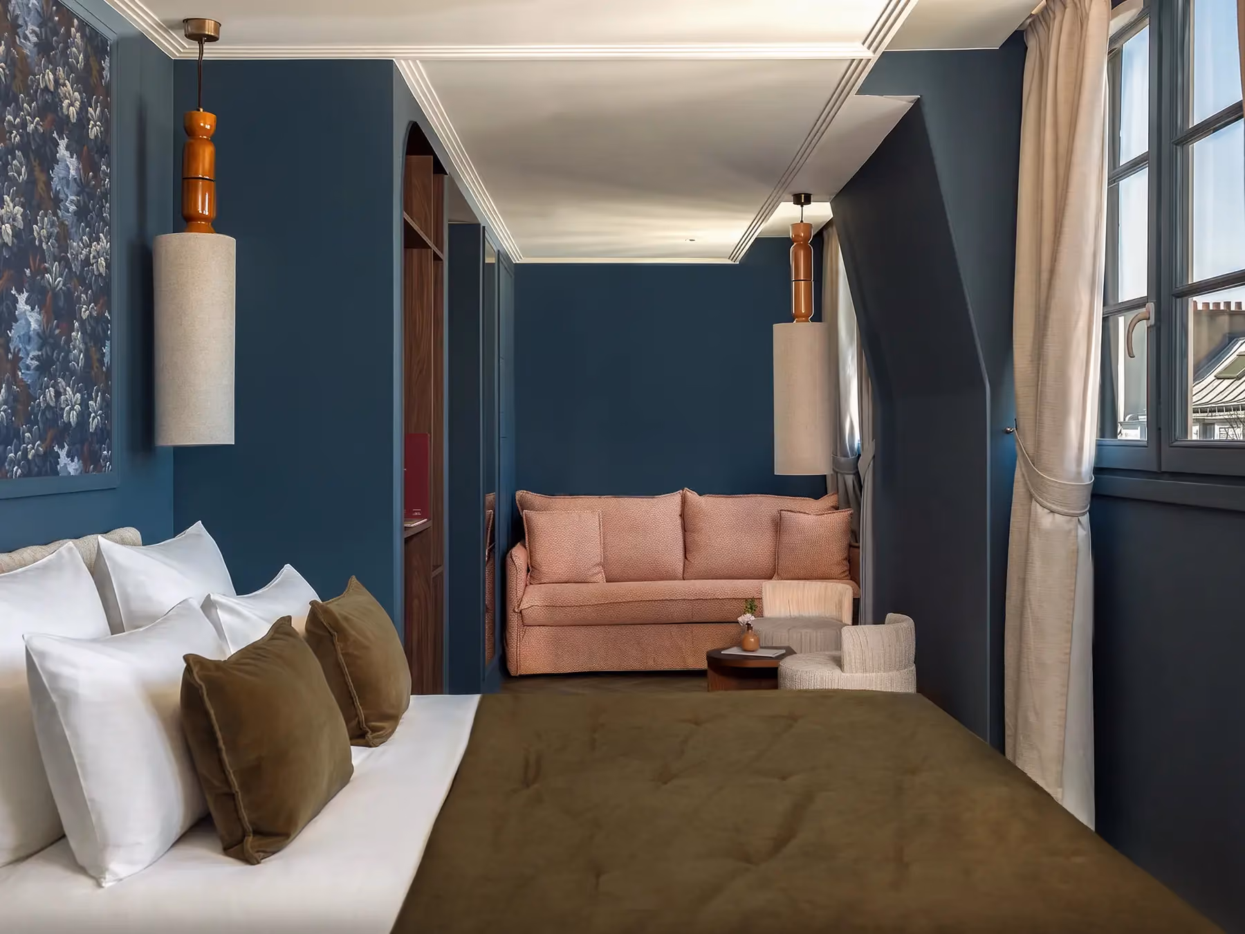 Chambre aux murs bleu canard avec canapé rose, lit à jeté olive et vue sur les toits parisiens.