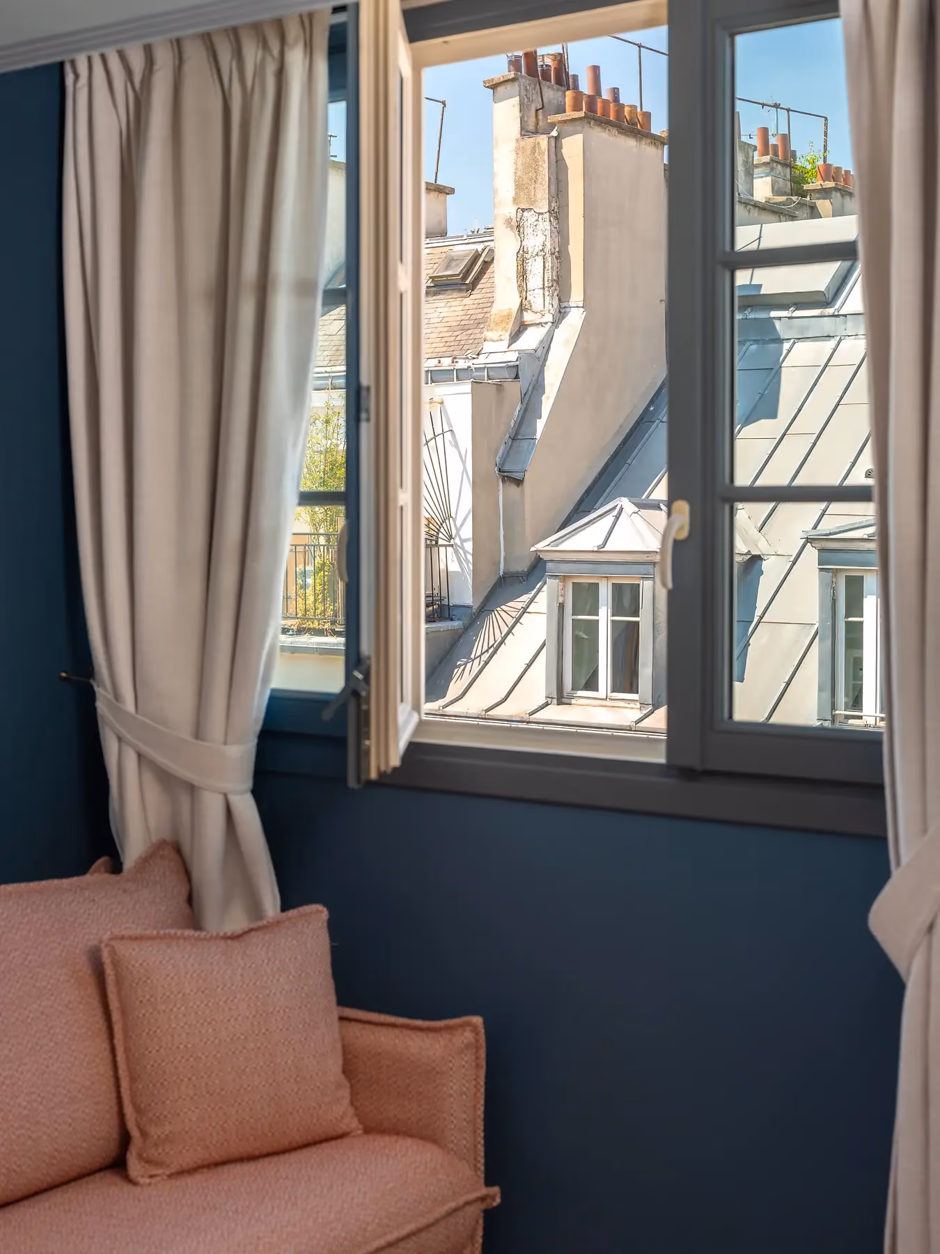 Fenêtre ouverte sur les toits de Paris, avec fauteuil rose et mur bleu marine.