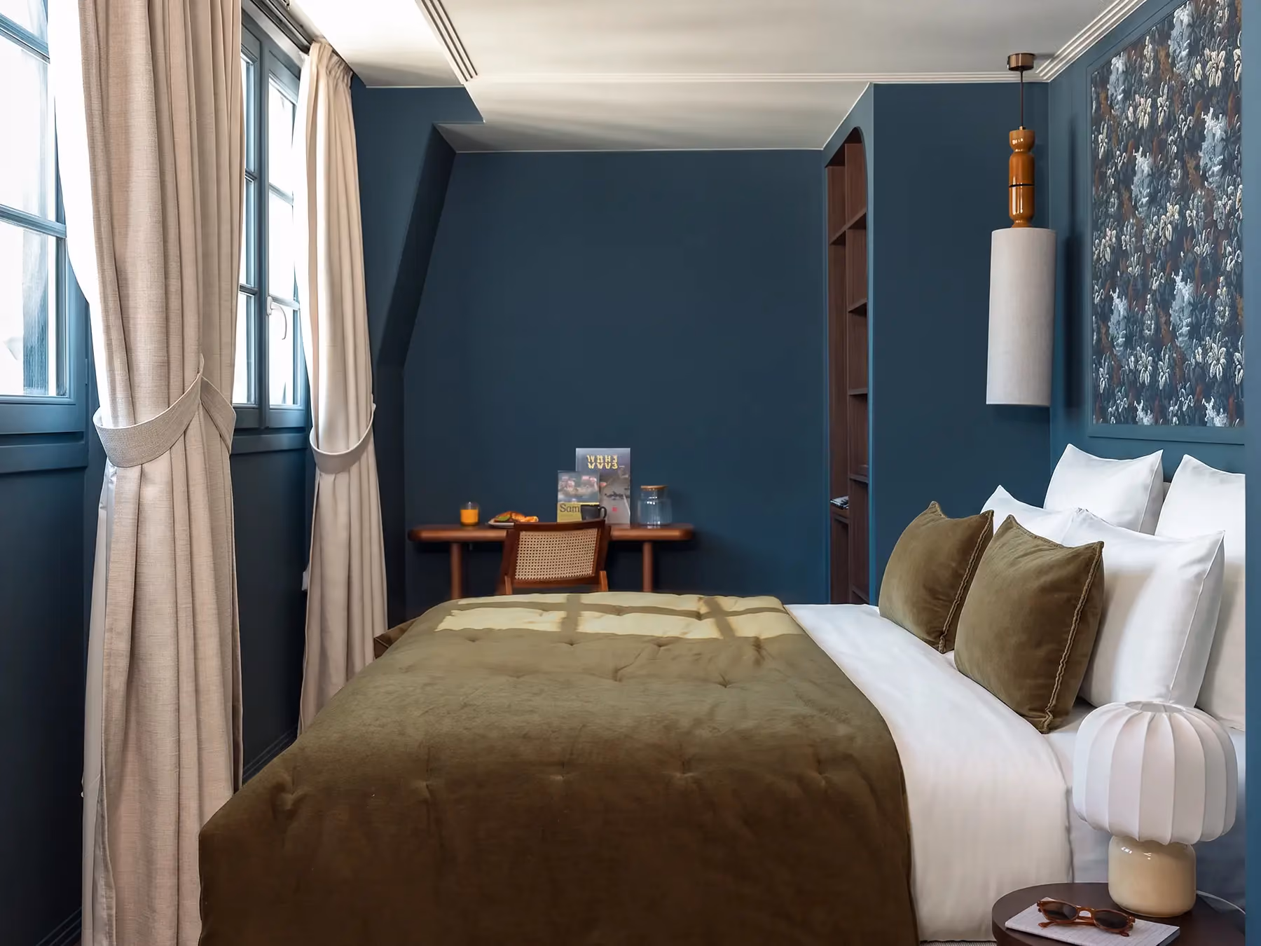 Chambre bleu canard avec lit à jeté olive et deux fenêtres à rideaux beiges.