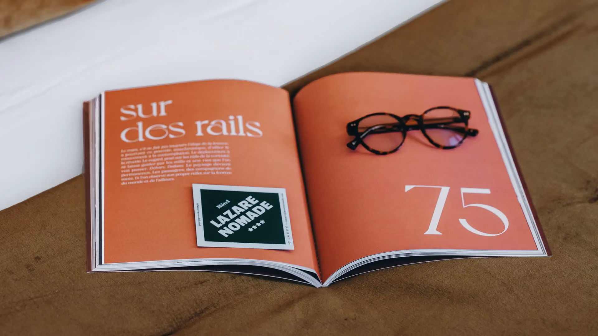 Magazine ouvert sur "sur dos rails" et "75" avec lunettes et carte "Hôtel Lazare Nomade", posé sur un lit.
