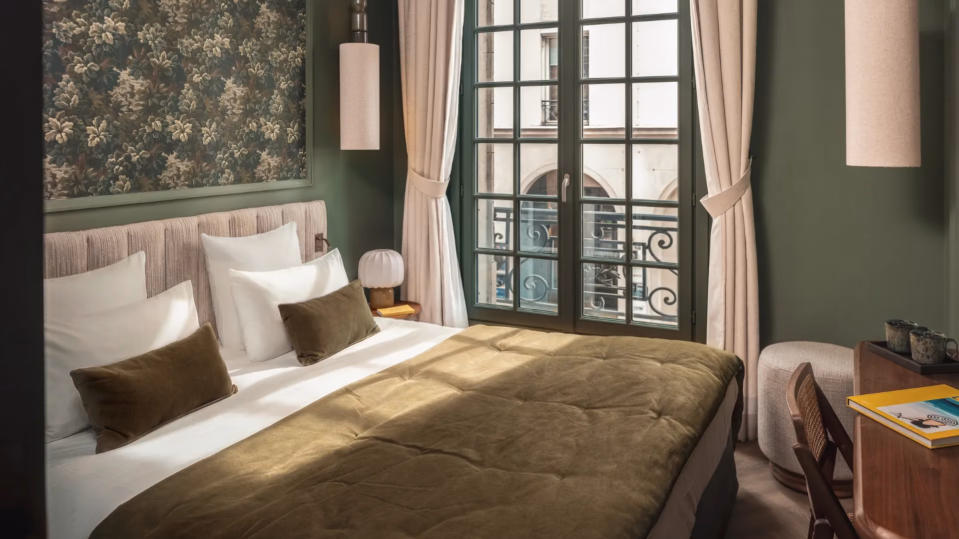 Chambre vert sauge avec lit couvert d'un jeté olive, tapisserie florale et fenêtre ouvragée sur balcon parisien.