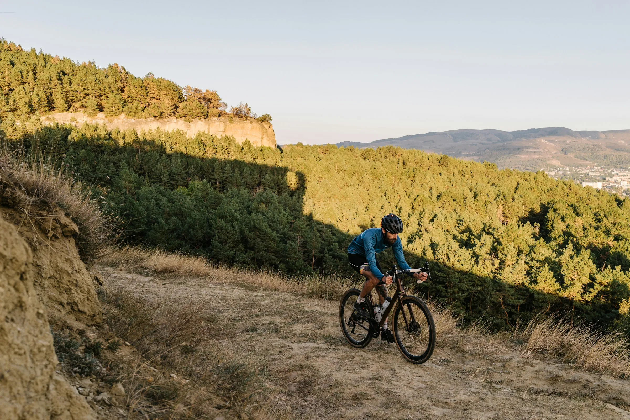 A cyclist climbing a gravel ridge track above a forested valley in late afternoon light