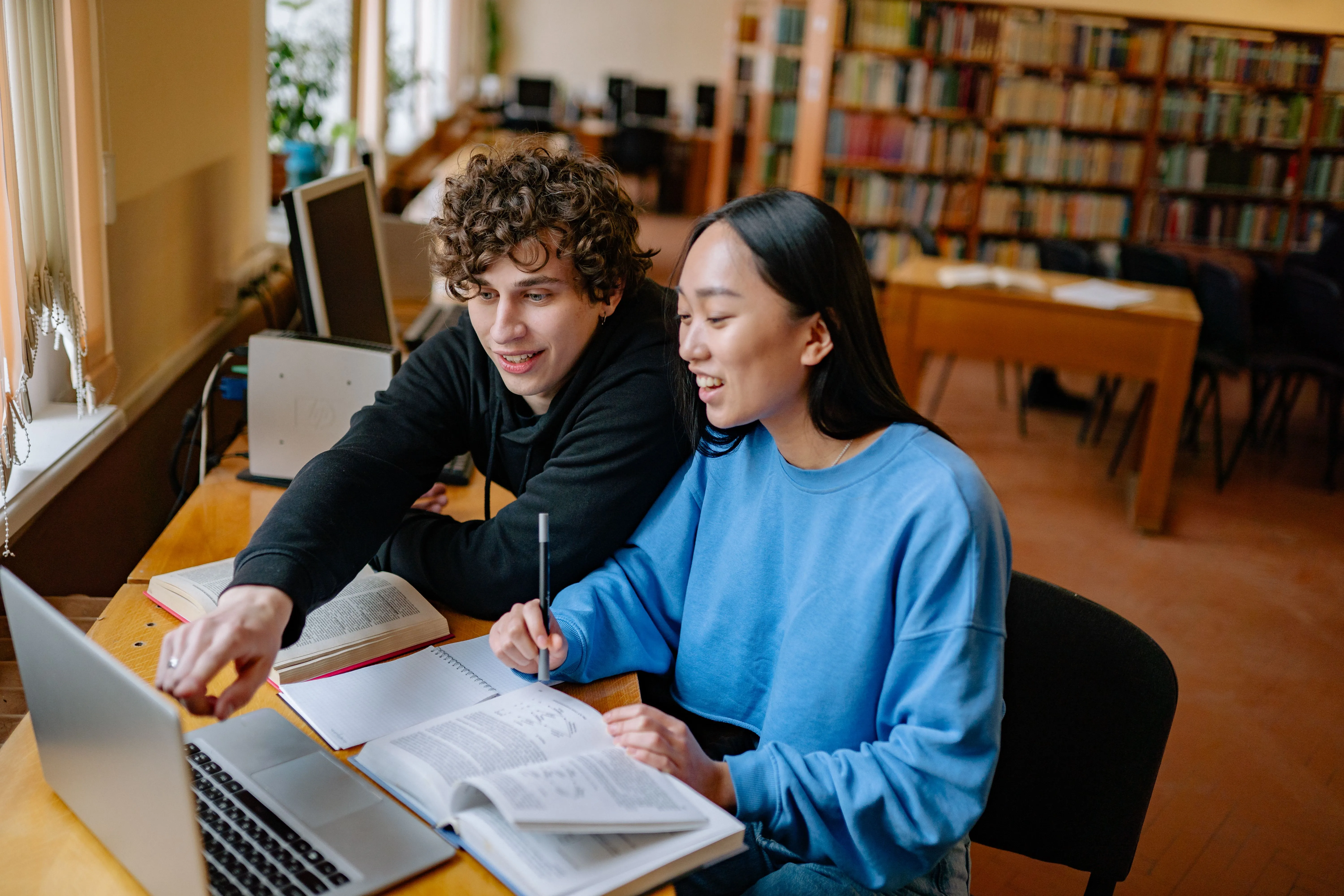 Two students working together on a laptop with books and notes