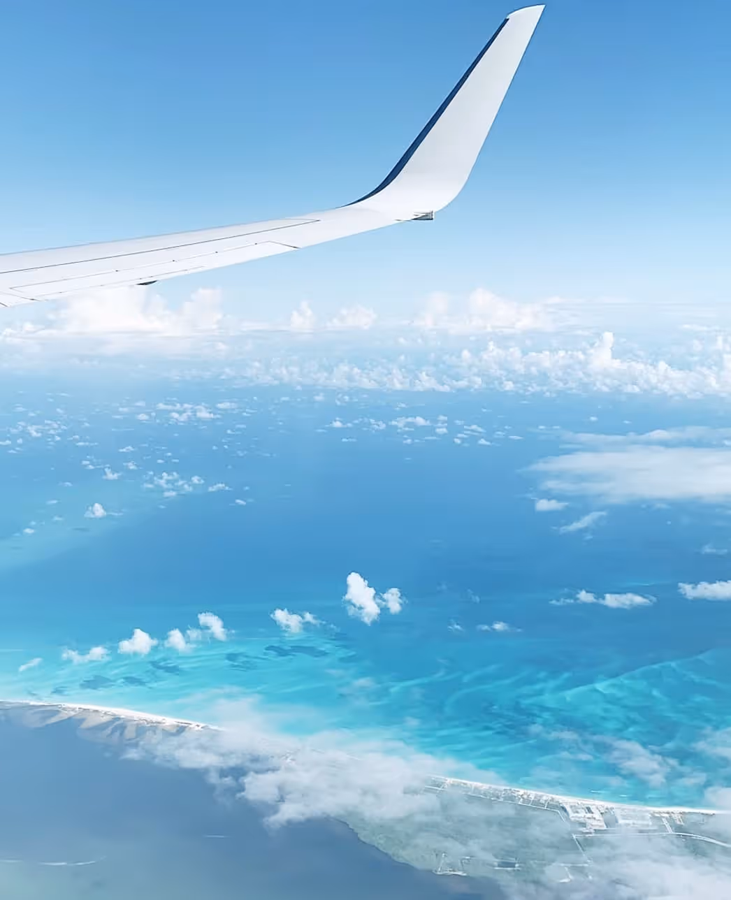 View of airplane wing over turquoise ocean with clouds and coastline below.