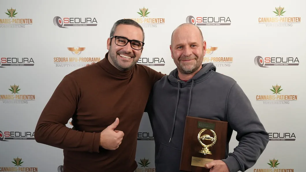 Zwei Männer lächeln, einer zeigt Daumen, der andere hält eine Plakette mit Goldauto darauf.