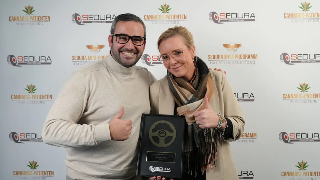 Zwei lächelnde Personen zeigen Daumen hoch und halten eine Trophäe vor einer Sedura-Wand.