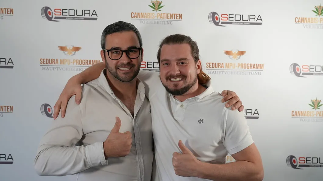 Zwei Männer lächeln, umarmen sich und zeigen Daumen hoch vor Werbewand von Sedura und Cannabis-Patienten.