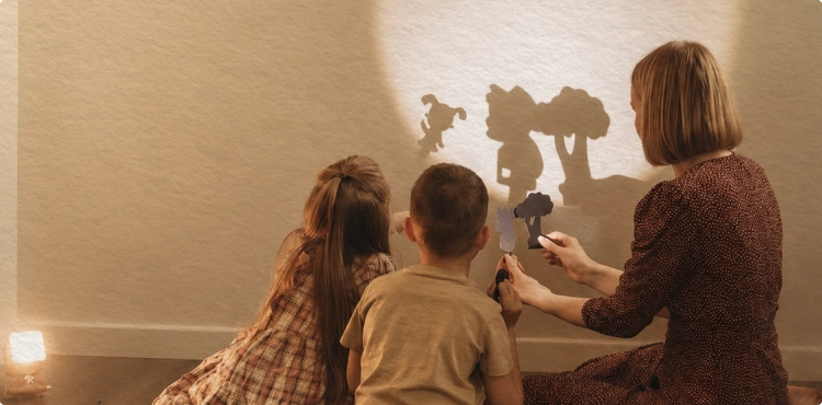 Woman and two children creating shadow puppets of a tree and animals on a wall.