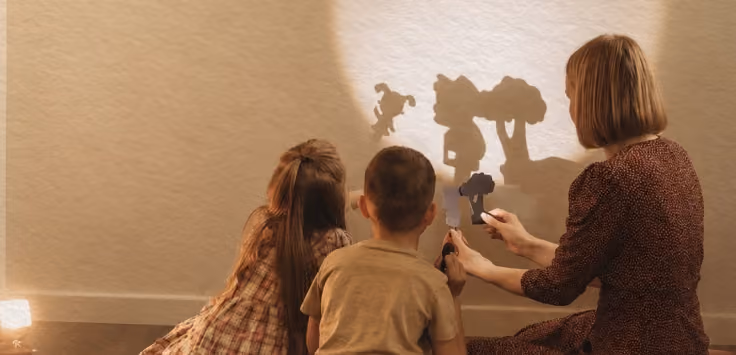 Woman and two children creating shadow puppets of a tree and animals on a wall.