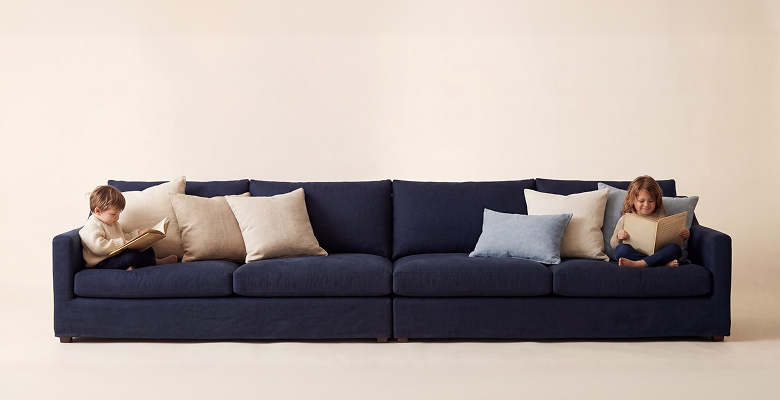 Two children sitting on opposite ends of a large navy blue sofa reading books with beige and blue pillows.