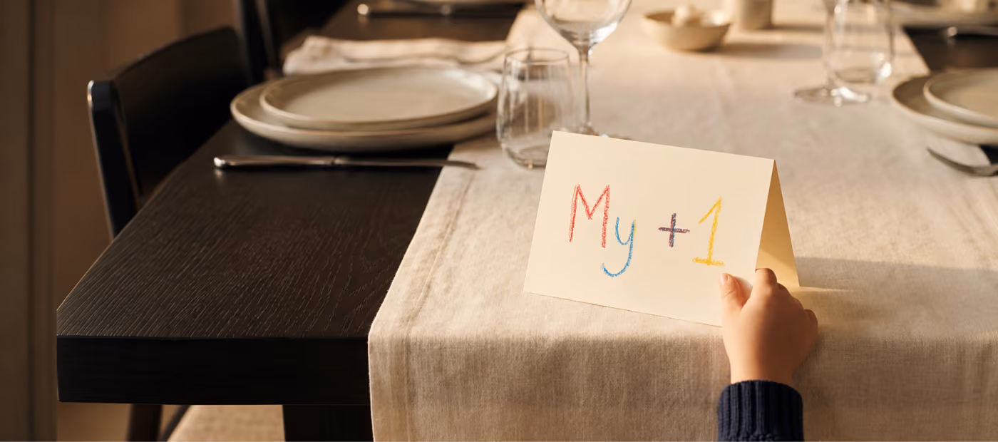 A child's hand holding a cream-colored folding card on a dining table with the colorful handwritten text 'My +1' on it, set on a beige tablecloth next to dinner plates, silverware, and glassware.