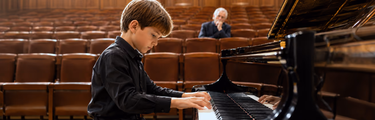 Young boy playing a grand piano in an empty concert hall while an older man watches attentively from the audience seats.