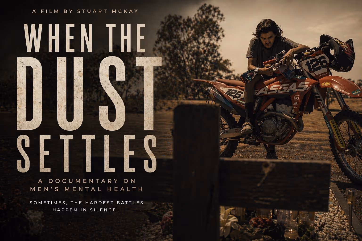 Young man sitting on a dirt bike near a grave marked by a wooden cross in a rural outdoor setting during sunset.