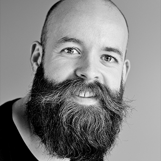 Smiling man with full beard and mustache, balding head, wearing a black shirt on a gray background.