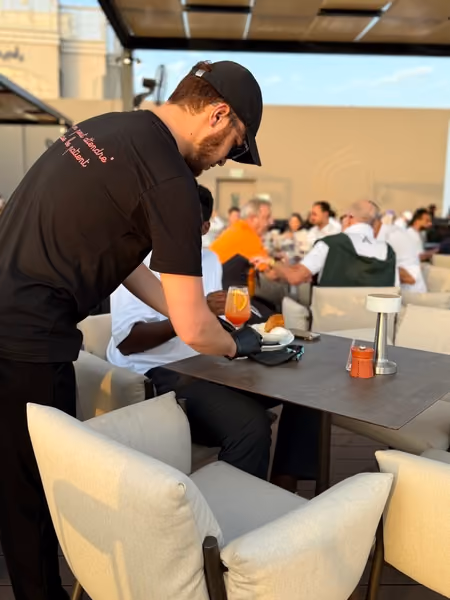Waiter setting plates at an outdoor table beneath palm trees at dusk