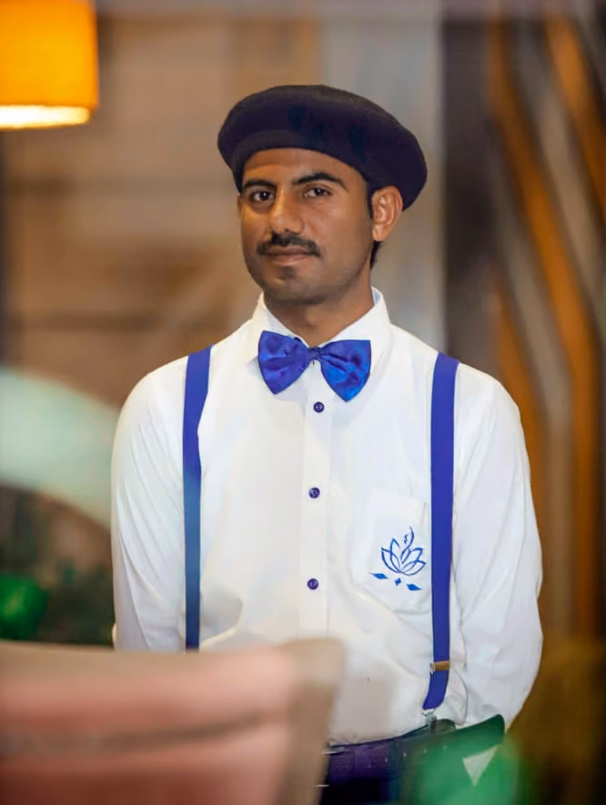 A man with a mustache wearing a black beret, white shirt with blue suspenders and a blue bow tie, standing indoors with a blurred background.