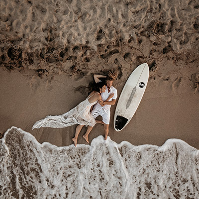 Drohnenaufnahme eines Hochzeitspaares, das mit einem Surfbrett an einem Strand von Mallorca liegt.