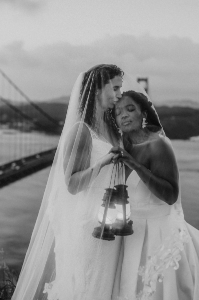 Artistic black and white portrait of LGBTQ newlyweds beneath a bridal veil holding a lantern with San Franciscos Golden Gate Bridge in the Background