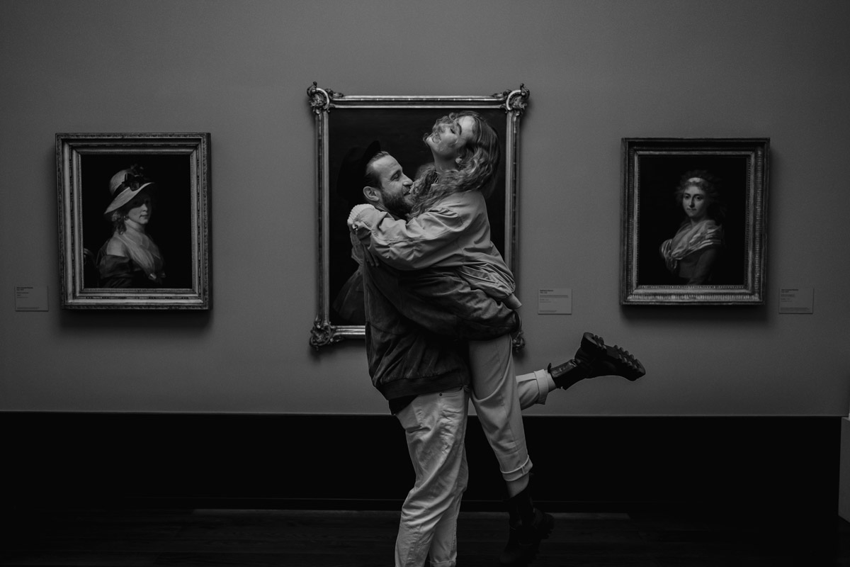 Zwischen Gemälden in einem Museum hebt ein Mann seine Freundin hoch. Schwarz-weiß Paarfoto.