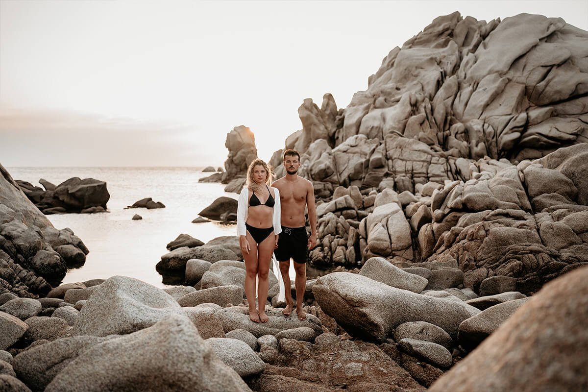 Ein in Bikini und Badehose bekleidetes Paar steht für ein Pärchen Shooting am felsigen Strand von Sardinien.