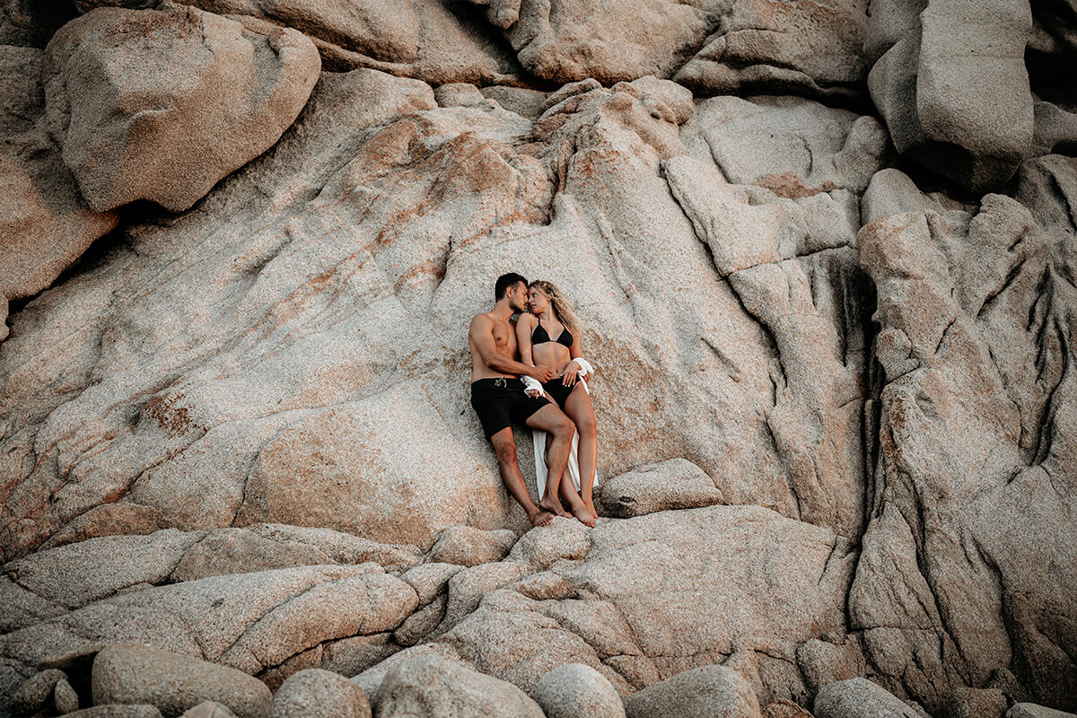 Ein verliebtes Paar steht in Badekleidung an eine Felswand auf Sardinien gelehnt und schaut sich an.