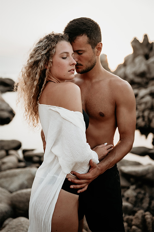 Ein in Bikini und Badehose bekleidetes Paar steht für ein Pärchen-Shooting am felsigen Strand von Sardinien.