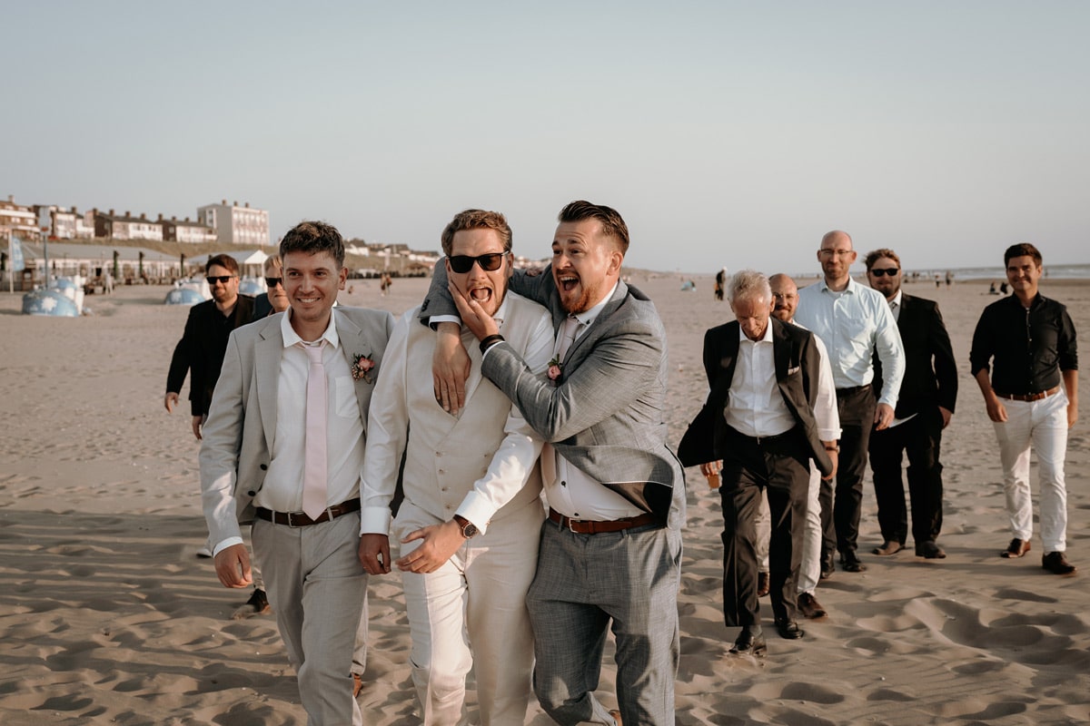 Ein Bräutigam geht mit seinen Freunden am Strand entlang und wird dabei von seinem Trauzeugen umarmt.