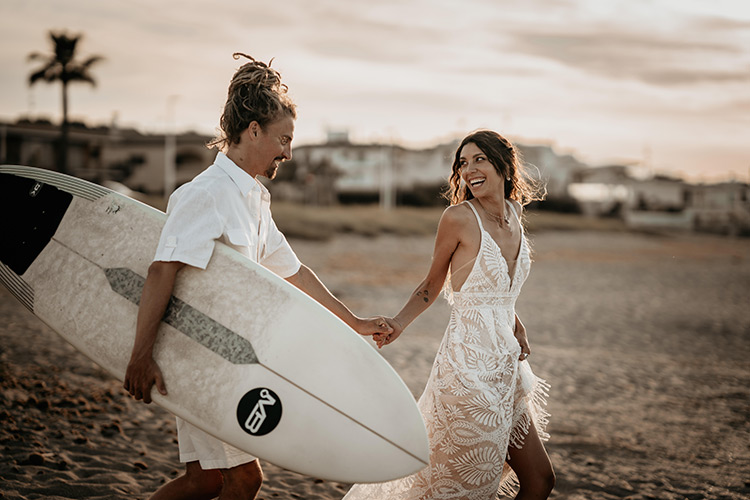 Ein Brautpaar läuft mit Surfbrett auf das Meer zu.