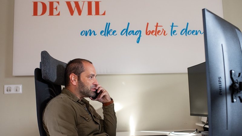 A Telsmart colleague at his desk in Kasterlee, under the painted wall text De wil om elke dag beter te doen