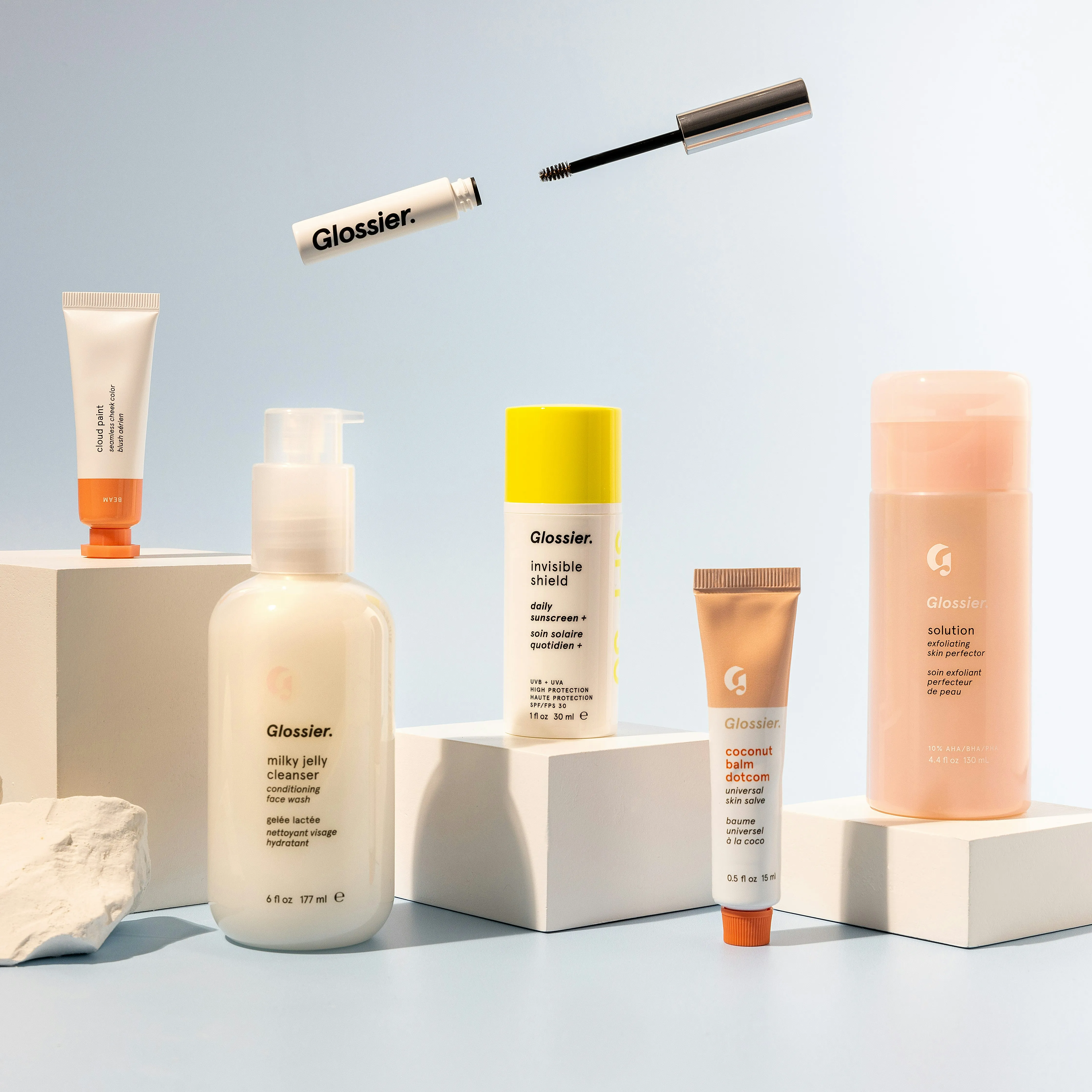 Zes cosmetische producten van Glossier op witte blokken tegen een lichtblauwe achtergrond.