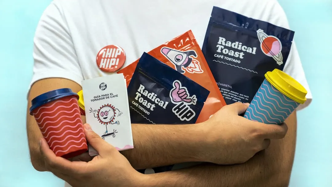 Persoon met witte t-shirt houdt kleurrijke koffiezakken, bekers en een gids voor koffiedrinkers vast.