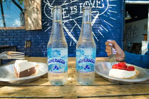 Twee flessen Mineragua bruiswater op tafel met stukken taart en een hand met vork.