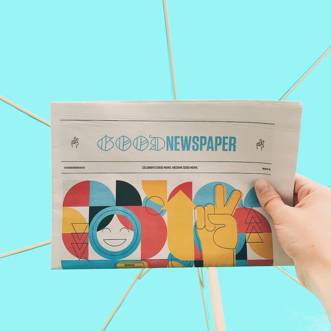 Hand houdt een krant vast met vrolijke illustraties en de titel Good Newspaper op een blauwe achtergrond.