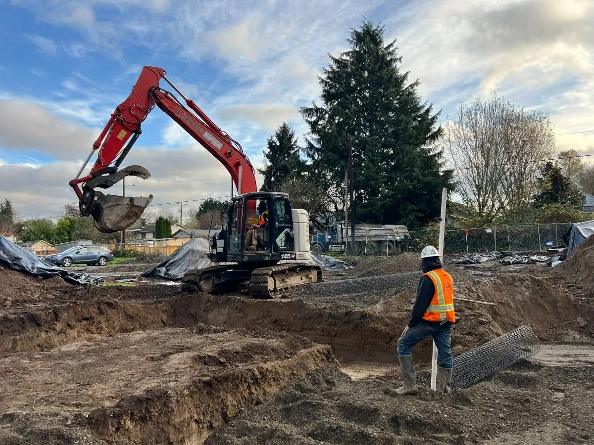 Excavator alongside open trenches as a contractor checks the work on a Seattle-area site