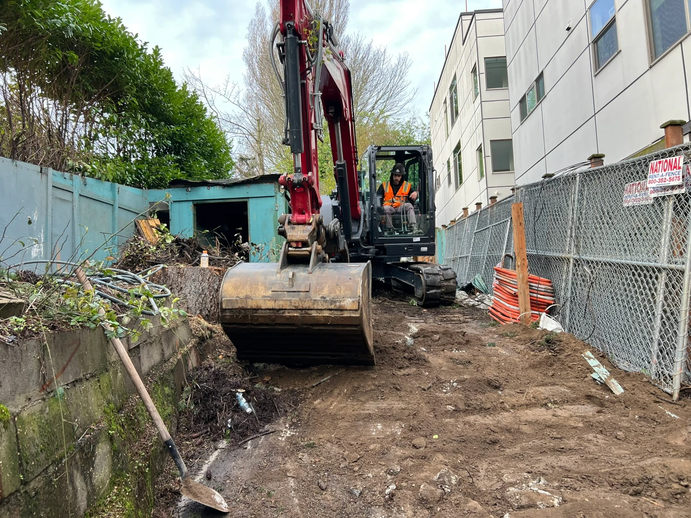 Operator maneuvering a compact excavator into a fenced backyard to begin structure demolition