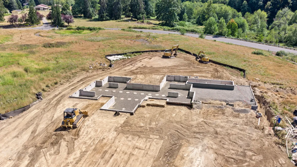 Aerial view of a completed residential concrete foundation with poured stem walls
and compacted gravel fill, with a dozer and two excavators finishing site grading.
