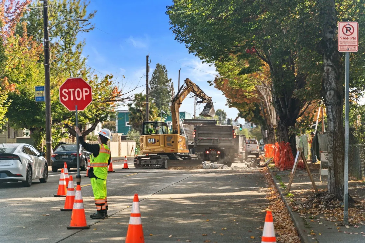 Construction worker in neon safety gear and helmet holding a stop sign directing traffic near an active roadwork site with a CAT excavator loading debris into a dump truck, surrounded by orange cones and trees with autumn leaves.