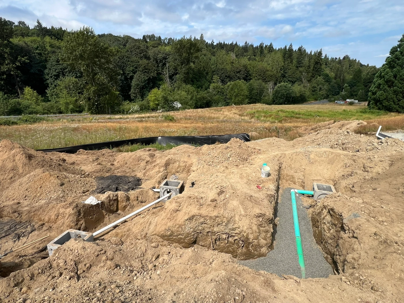 Storm line trench with green pipe on gravel bedding running between two concrete catch basins