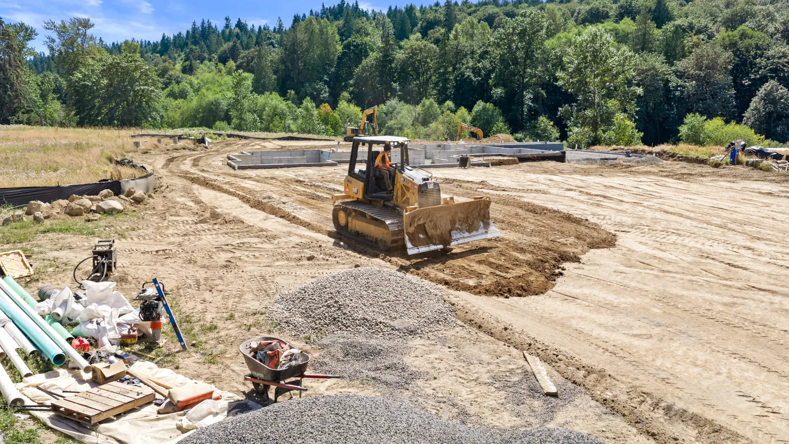 Operator running a CAT dozer to spread and grade fill dirt near a concrete foundation, with pipe and gravel stockpiles in the foreground