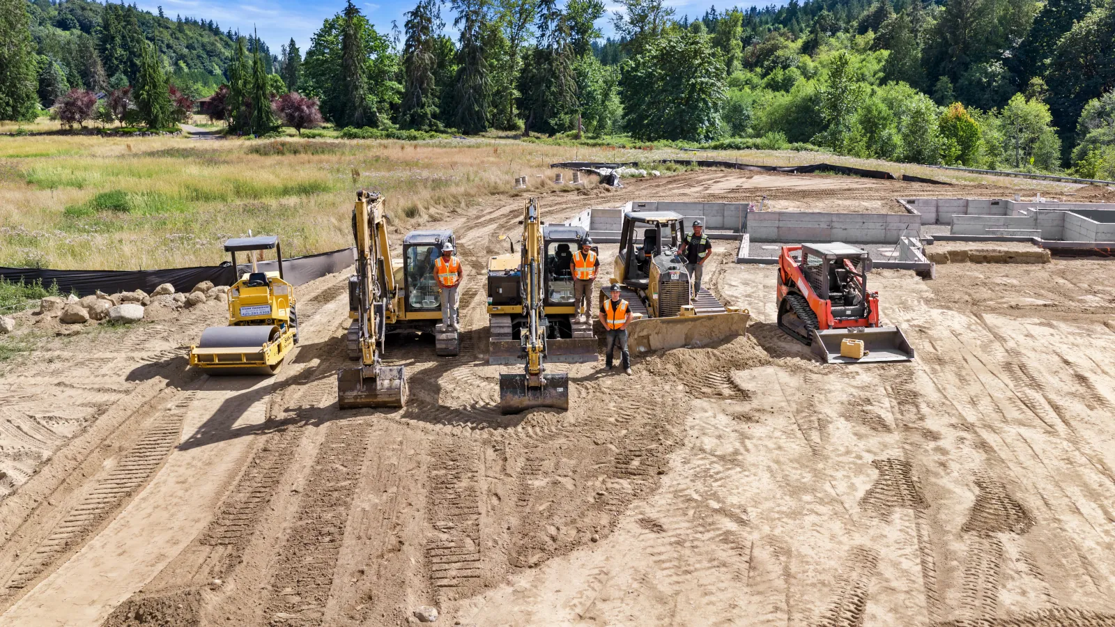 Drone overview of a new home foundation, graded pad, and silt fencing with excavators working the far end of the lot