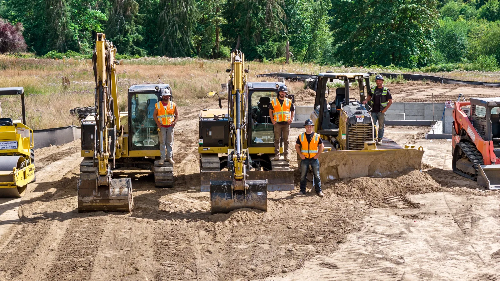 	Close view of an excavation crew posed with their machines on a compacted dirt pad next to a concrete foundation